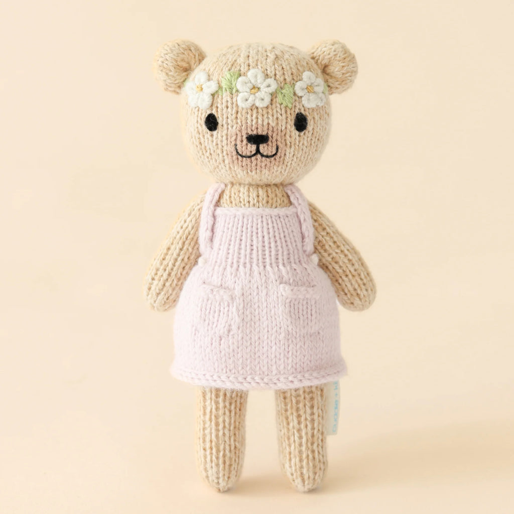 Cuddle + Kind Tiny Olivia the Honey Bear wears a light lavender dress with pockets against a pale background. This hypoallergenic, handmade toy features a simple smile, black eyes, and a crown of small white flowers on its head.
