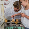 Two children enjoy a Wooden Tea Set on a vintage perforated table. One places a toy in a hole while the other admires the rustic setting, sipping almond chai, creating an ambiance of playful nostalgia and warmth.