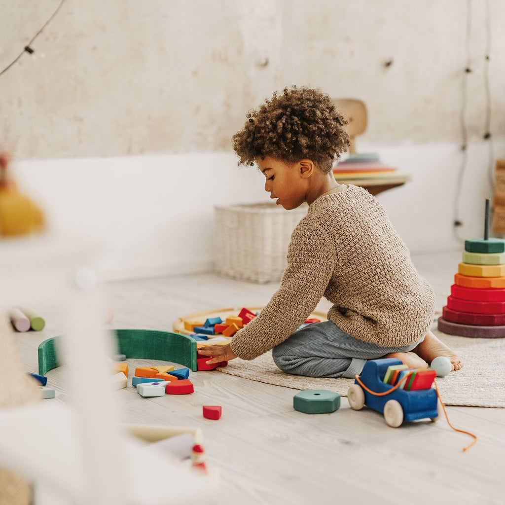A child with curly hair, wearing a brown sweater and jeans, joyfully plays on the floor with Grimm's Pull Along Truck and other wooden blocks. The warmly lit room is cozily decorated in a minimalist style, and all toys are non-toxic for safe playtime adventures.