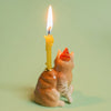A ceramic figurine similar to the Maine Coon Cat Cake Topper, featuring a party hat and whimsical candle holder design, holds a lit yellow candle against a soft green background—ideal for cat-themed events.