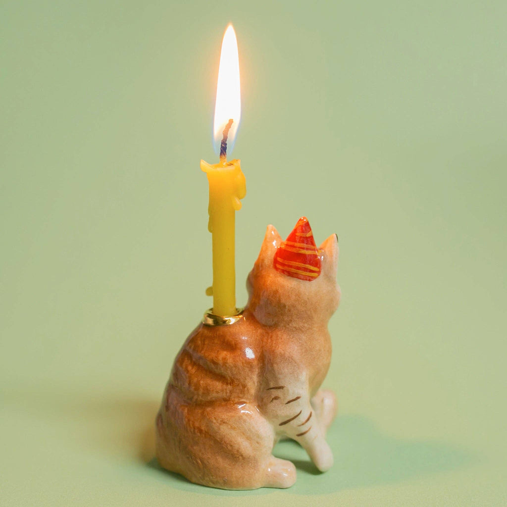 A ceramic figurine similar to the Maine Coon Cat Cake Topper, featuring a party hat and whimsical candle holder design, holds a lit yellow candle against a soft green background—ideal for cat-themed events.