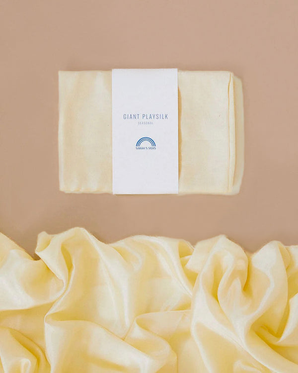 Folded and loose yellow Sarah's Silk Giant Lambswool Playsilk set against a beige background. Crafted from pure mulberry silk, the folded piece has a label that reads "Giant Playsilk." The fabric's smooth and soft texture enhances its Winter colorways appeal.