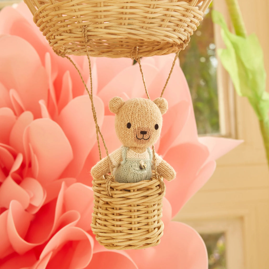 A Cuddle + Kind Tiny Charlie the Honey Bear, wearing overalls, sits inside a hot air balloon-like wicker basket. It's stuffed with hypoallergenic polyfill and surrounded by a large pink paper flower, with sunlight filtering through the window in the background.
