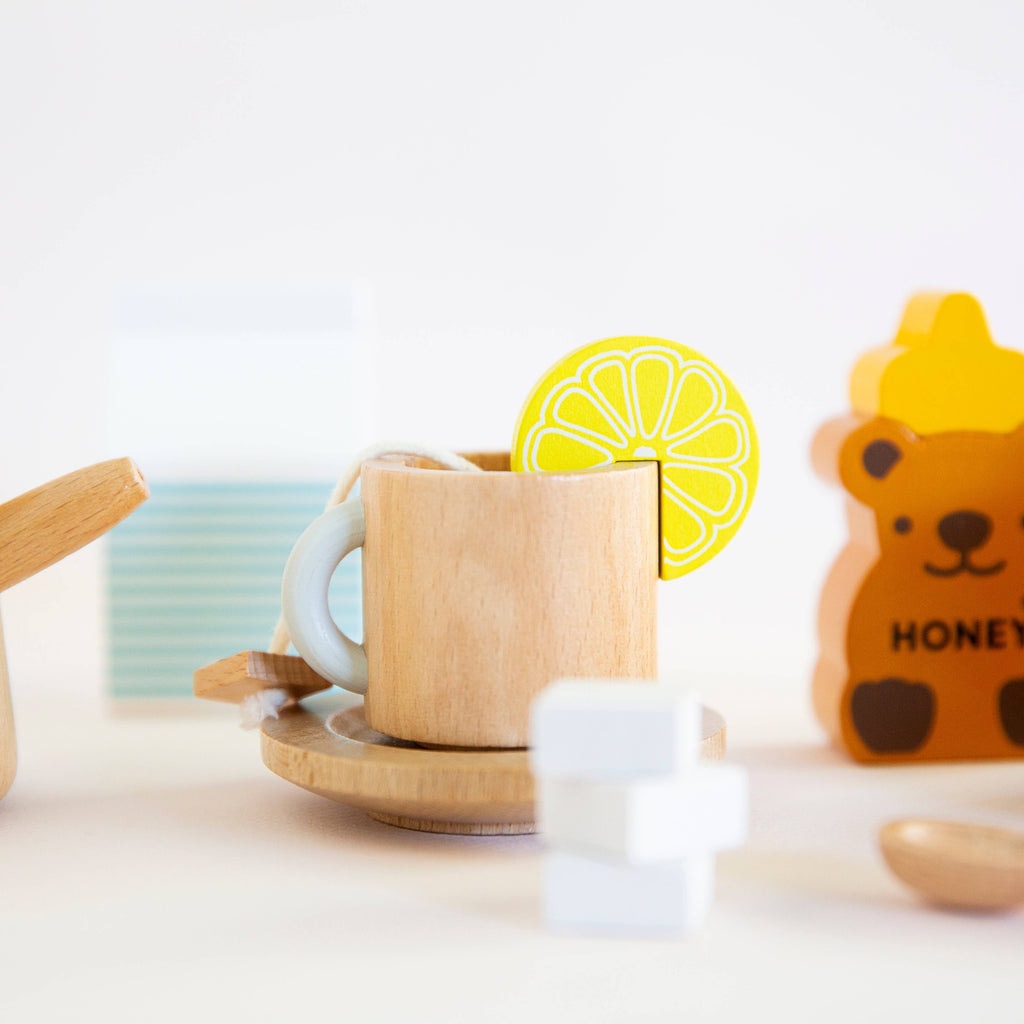 The Wooden Tea Set includes a wooden cup with a lemon slice, a dish, a white sugar cube, and a bear figure labeled "honey," creating an inviting and playful scene against a minimalist background.