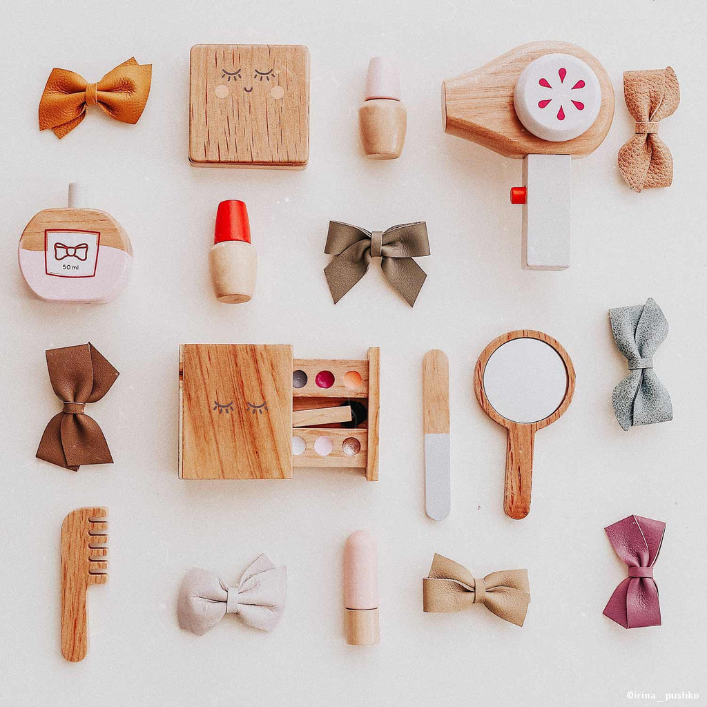 The Wooden Beauty Kit, a timeless toy, includes wooden makeup items like a hairdryer, compact, lipsticks, mirror, and comb with non-toxic paint. It also features five colorful bows and is beautifully arranged on a light background.