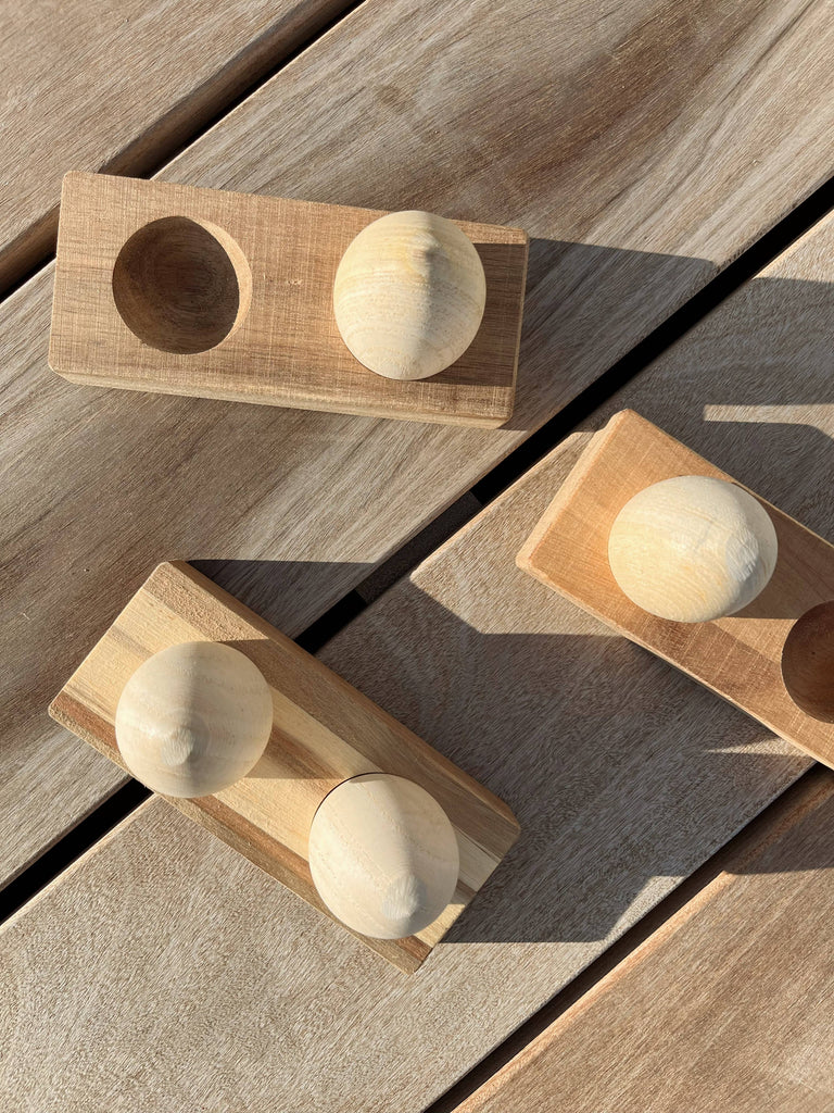 Three Wooden Egg Shakers with Stand rest on a wooden surface, casting gentle shadows in the sunlight. Perfect for encouraging developmental milestones, each shaker features a natural wood finish and is displayed on its sleek stand.