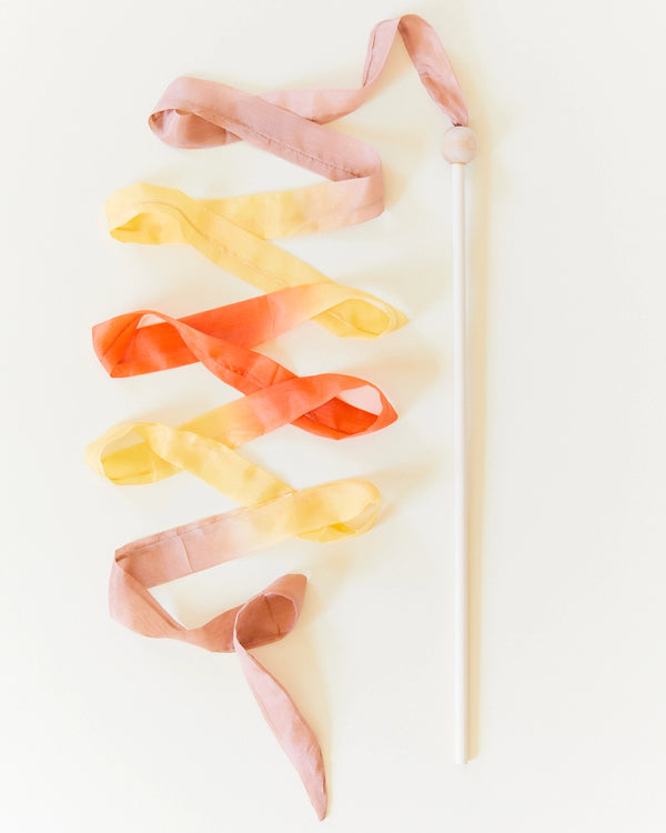 A white ribbon dance stick with a vibrant multicolored Sarah's Silk Boho Streamer Wand, perfect for encouraging open-ended play. The streamer transitions through shades of yellow, orange, and pink as it spirals down from the round knob at the top of the stick, enhancing gross motor skills against a white background.