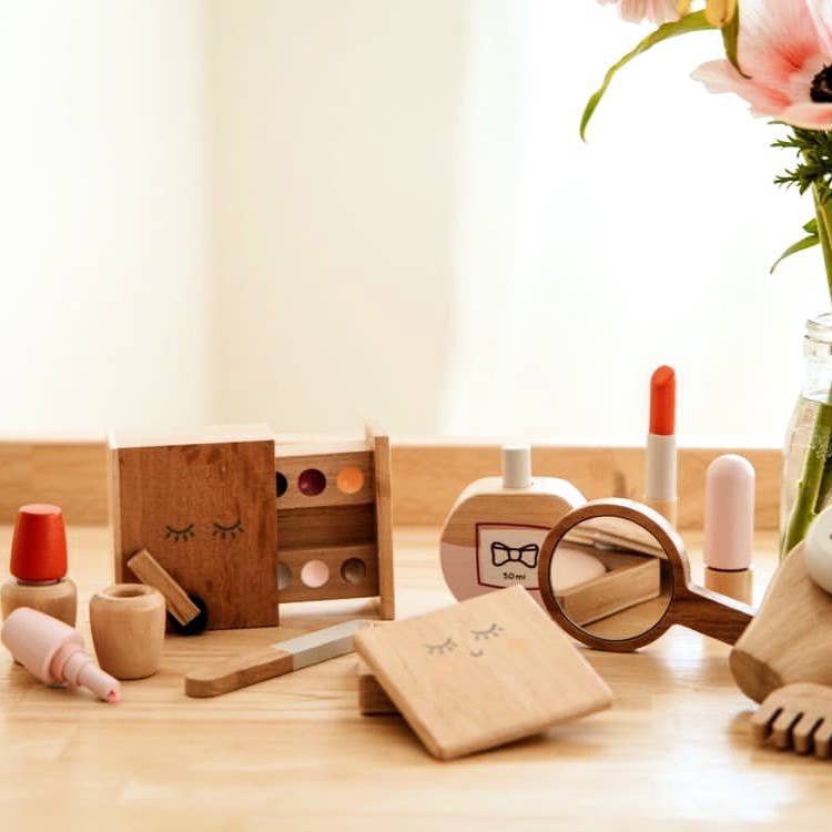 The Wooden Beauty Kit sits elegantly on the table, featuring a lipstick, eye shadow palette, mirror, and nail polish. Made with non-toxic paint and cute facial expressions, it's an enchanting playset. A vase of pink flowers adds charm to the corner.