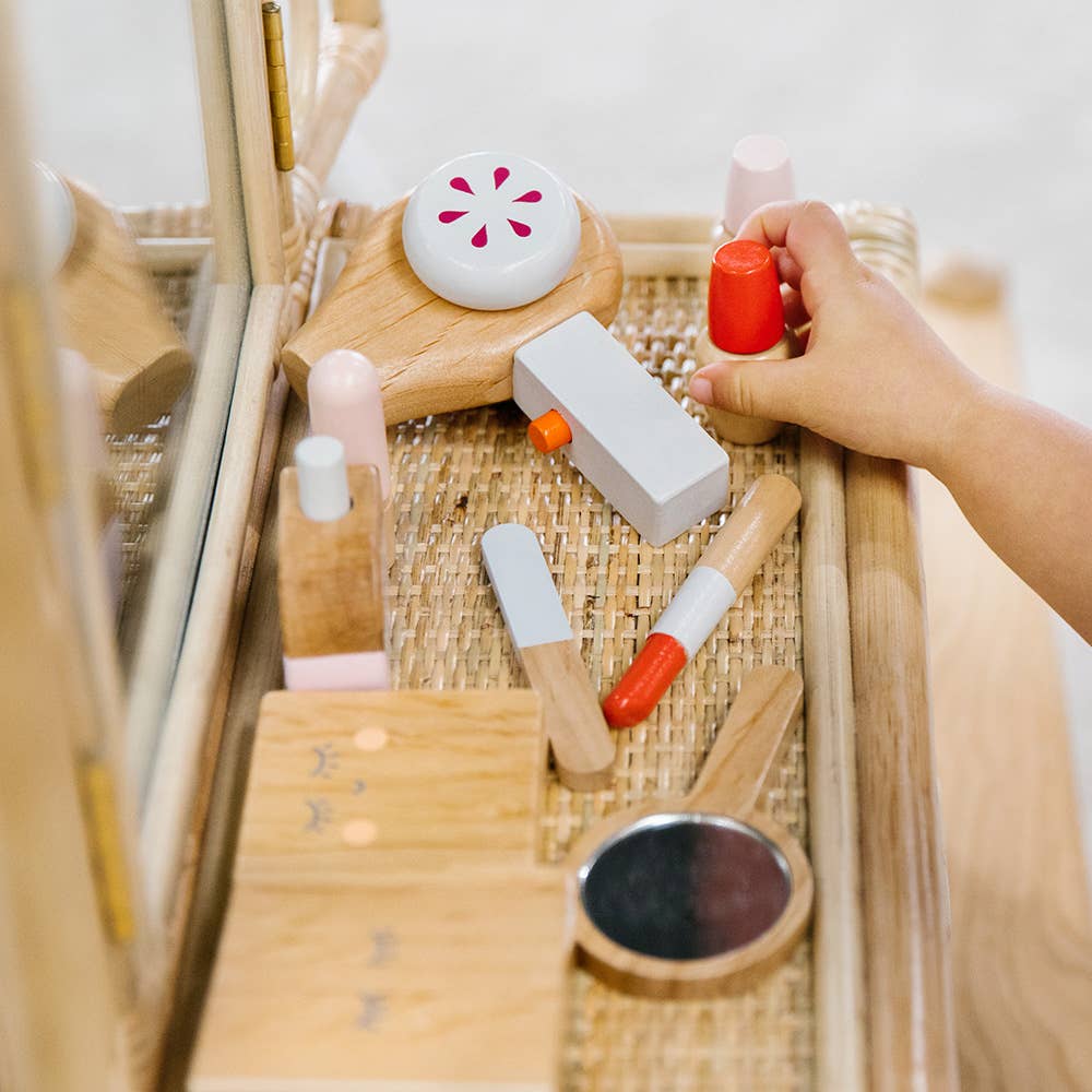 A child's hand grips a red toy bottle from the Wooden Beauty Kit on a wicker vanity table. The set boasts non-toxic painted wooden toys, including lipsticks, a compact, and a mirror, artfully displayed and perfectly mirrored in the timeless toy's reflection.