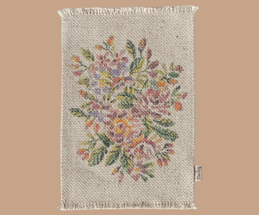 The Maileg Nursery Set includes a rectangular woven mat with fringed edges, decorated in a colorful floral pattern on a cream background and finished with a small fabric tag on the lower right edge. Ideal for placing under decor pieces.