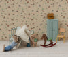In a playful scene, a charming mouse doll in a striped shirt relaxes on a cushion, surrounded by the Maileg Mouse Rocking Horse, a blue cupboard, yellow chair, clock, and miniature accessories, all set against patterned wallpaper.