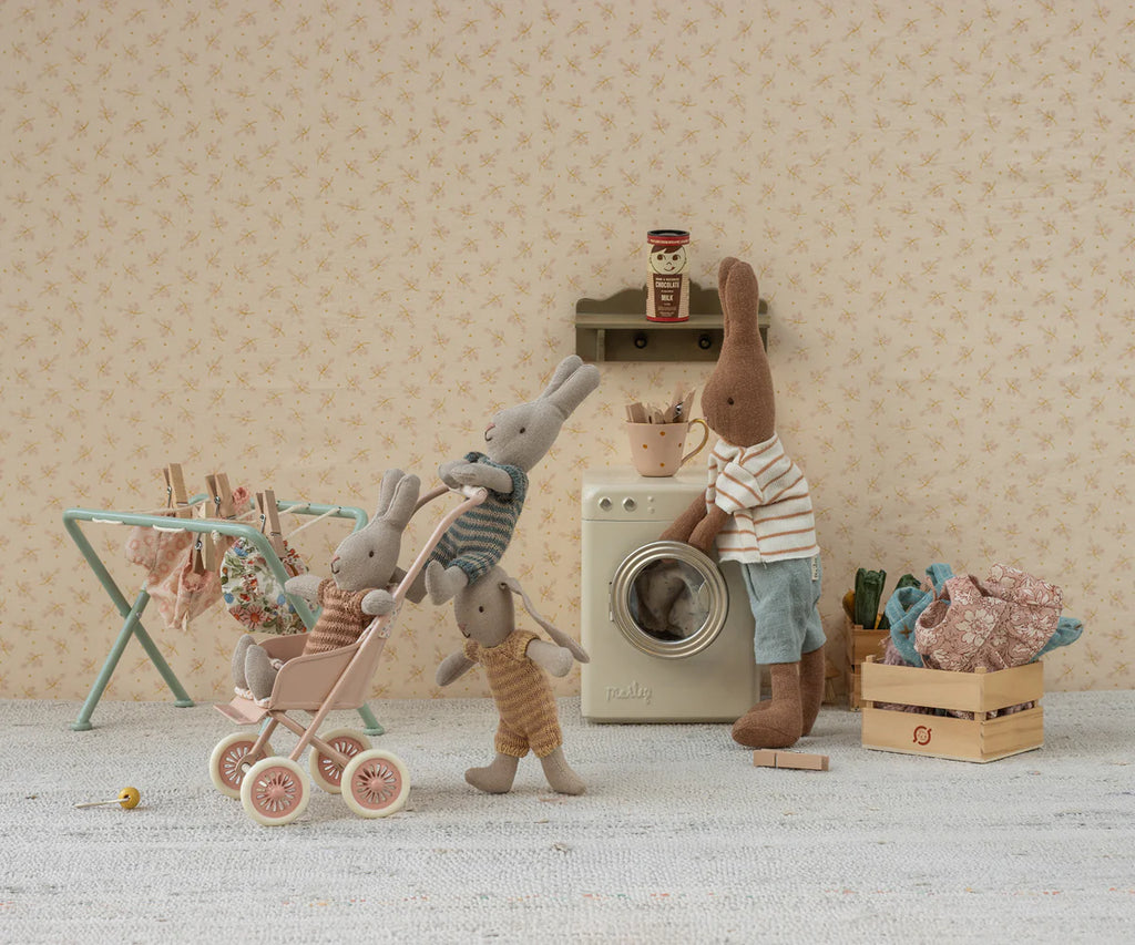 Four plush rabbits are in a vintage laundry room. One pushes two little ones in the Maileg Stroller, Micro - Powder, while another stands by a toy washing machine. Clothes hang on a drying rack nearby.