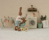 A stuffed bunny in a striped shirt and blue shorts pushes a smaller bunny in the Maileg Stroller, Micro - Mint. Mini laundry hangs on a rack, with a toy washing machine and plants adding to the cozy scene.