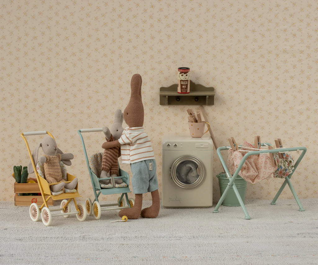 Two stuffed rabbits do laundry in a micro, vintage-style room. One rabbit pushes a Maileg Stroller, Micro - Yellow with a smaller bunny inside, while laundry dries on a rack and a toy washing machine adds to the cozy, whimsical scene.