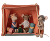 The Maileg Happy Camper Tent - Rust includes three plush mice: two are seated inside with rolled-up sleeping bags, dressed in colorful outfits and hats, while another stands outside holding the flap. The playful camping scene is completed with scattered wooden sticks.