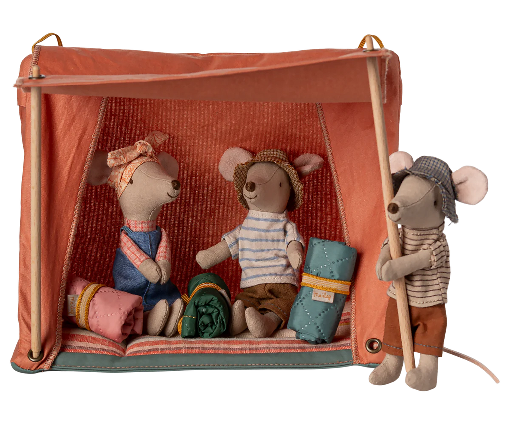 The Maileg Happy Camper Tent - Rust includes three plush mice: two are seated inside with rolled-up sleeping bags, dressed in colorful outfits and hats, while another stands outside holding the flap. The playful camping scene is completed with scattered wooden sticks.