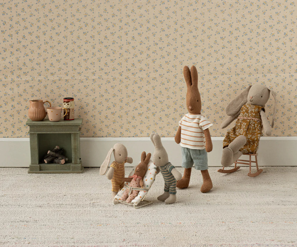 Five pastel-dressed stuffed rabbits are in a cozy room with floral wallpaper. One sits in the Maileg My Babysitter - Multi Dots chair, three smaller ones play together, and one stands nearby. A green shelf with pitchers and logs is behind them.