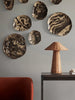 A modern room features a dark gray wall adorned with abstract, swirled-pattern metallic plates in different sizes, accompanied by a sleek Ferm Living Dou Table Lamp - Natural on a dark side table beside a red armchair.