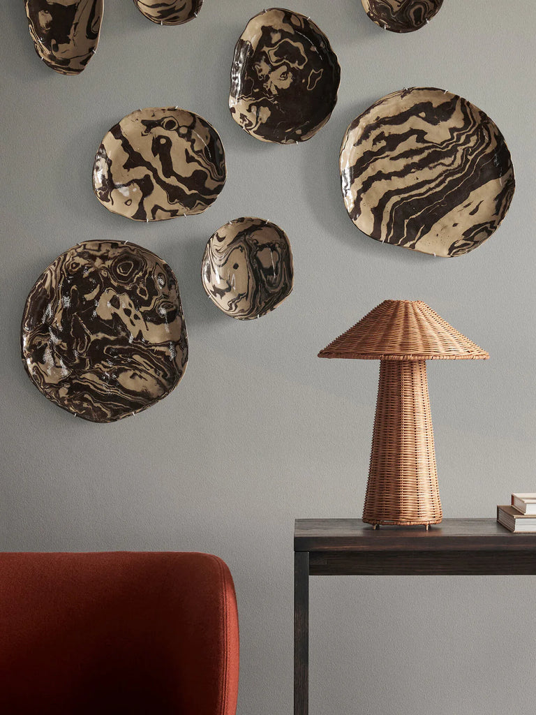 A modern room features a dark gray wall adorned with abstract, swirled-pattern metallic plates in different sizes, accompanied by a sleek Ferm Living Dou Table Lamp - Natural on a dark side table beside a red armchair.