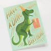 The Dino-mite Birthday Card shows a green dinosaur in an orange party hat holding a gift with "Have a dino-mite birthday" in playful letters. It includes a metallic gold envelope and has a blank interior for your message.