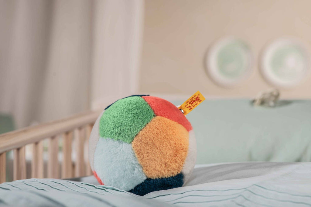 A Steiff Multicolored Plush Ball with Rattle and yellow tag rests on a light blue-striped bed in a softly lit bedroom. Easy to grip, this plush ball is near a wooden crib and two decorative plates in the background.