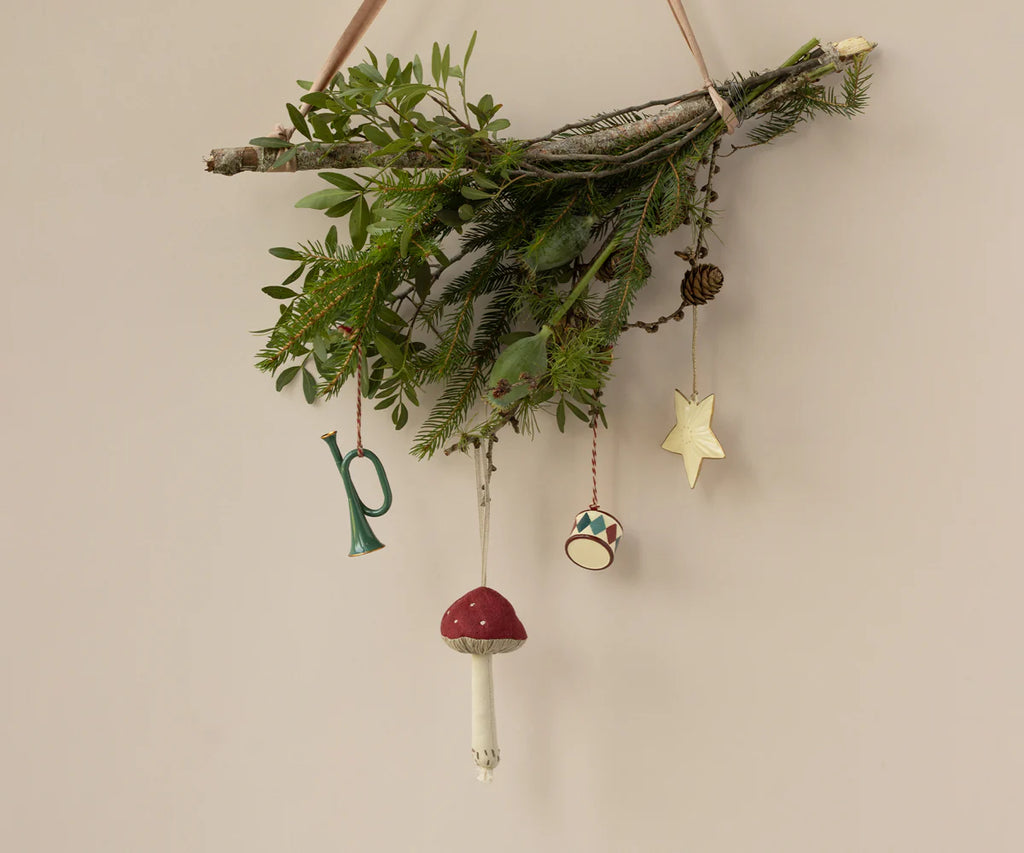 A decorative wall arrangement of twigs and greenery features metal ornaments with handpainted details, including the Maileg Metal Ornament Trumpet in Dark Green. This charming piece, along with red and white mushrooms, a drum, and a gold star, brings a festive spirit reminiscent of Christmas tree decorations.