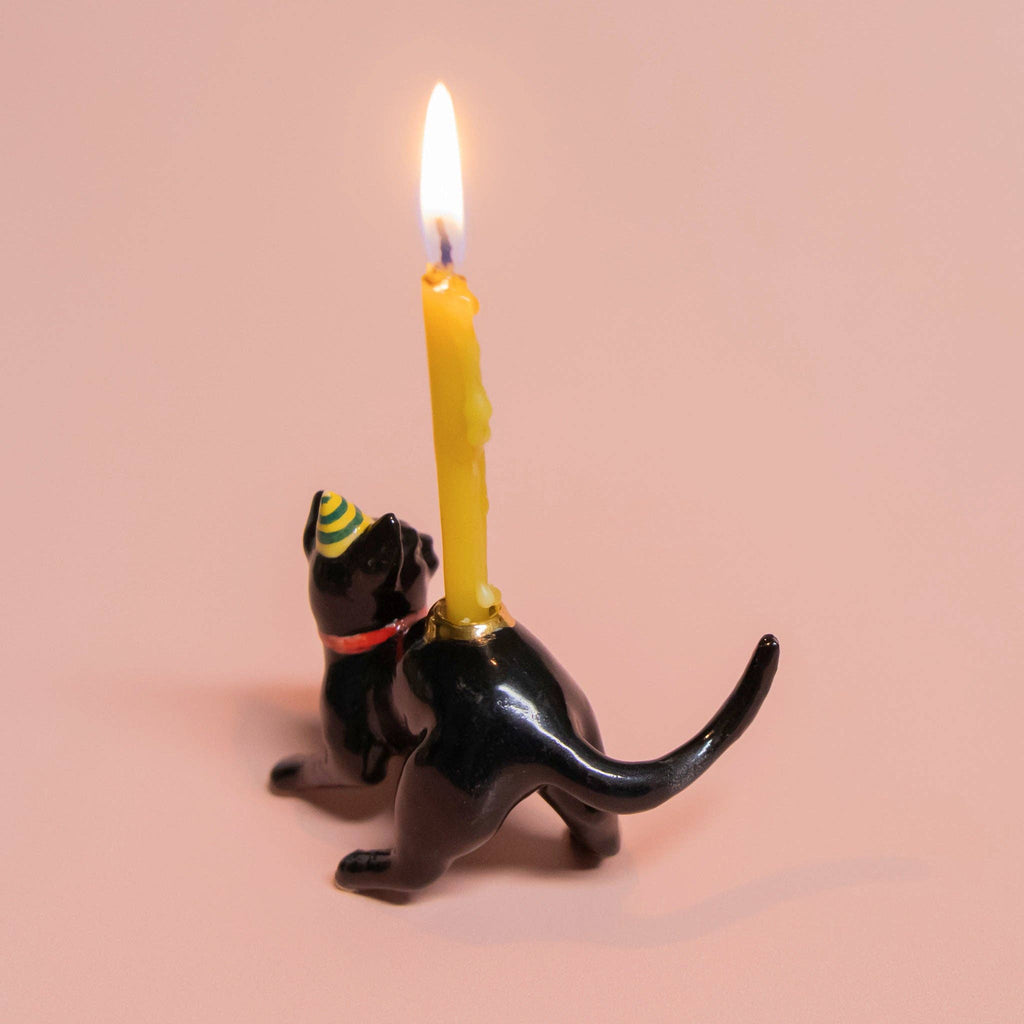 This Black Cat Cake Topper, ideal for cat-themed parties, showcases a black cat in a party hat and red collar with lifelike details. It holds a lit yellow candle on its back against a soft peach background.