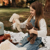 A young girl with long brown hair sits on a white blanket outdoors, holding a stuffed brown teddy bear and Harey the Bunny. She smiles in denim overalls next to a vintage-look suitcase on the blanket.