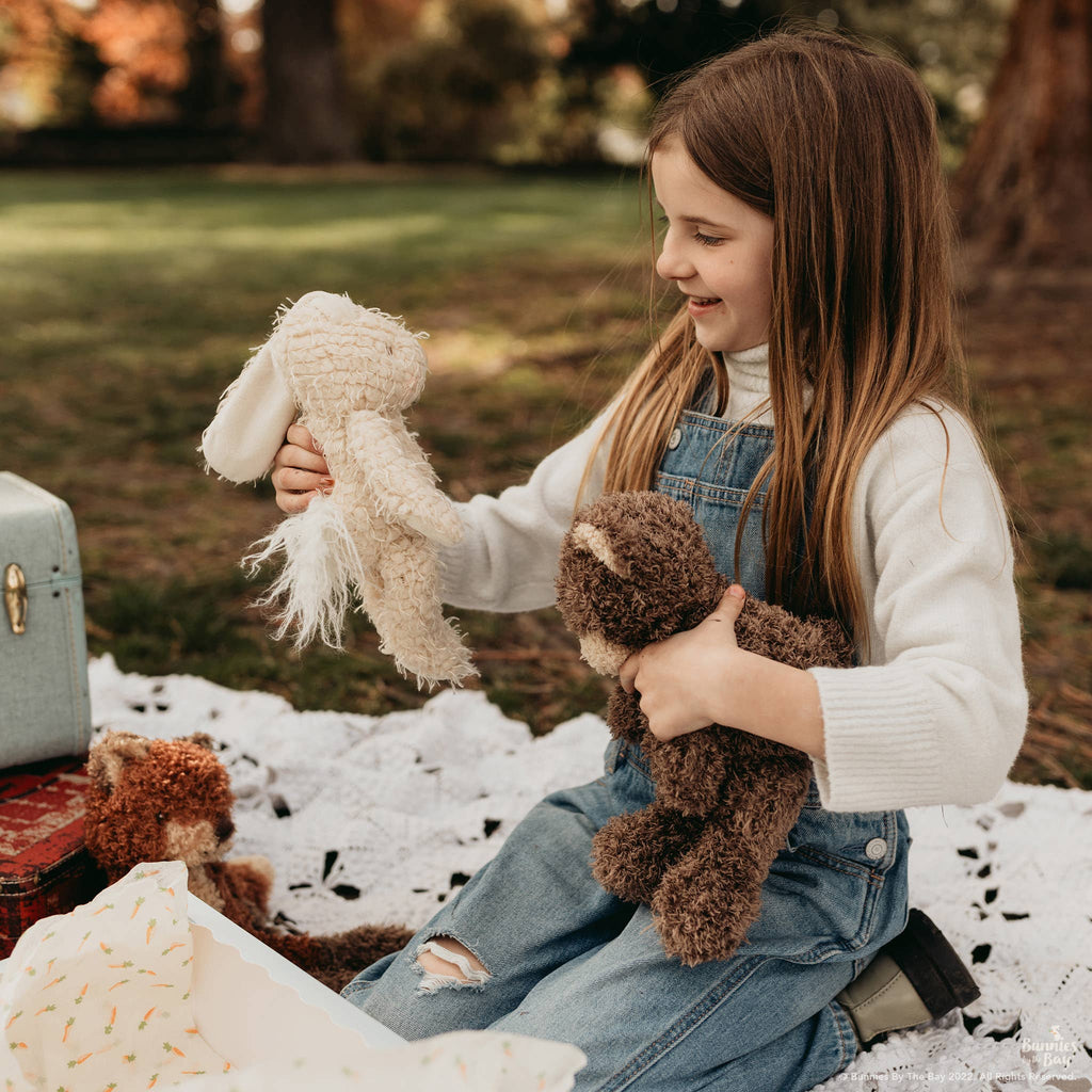 A young girl with long brown hair sits on a white blanket outdoors, holding a stuffed brown teddy bear and Harey the Bunny. She smiles in denim overalls next to a vintage-look suitcase on the blanket.