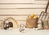 Three pocket-sized Poppet The Baby Hedgehog plush toys sit near a small wooden house labeled "Hedgehogs Welcome." A brown paper bag with potatoes and a garden fork, ideal for gardening, rest nearby against a light wooden wall.