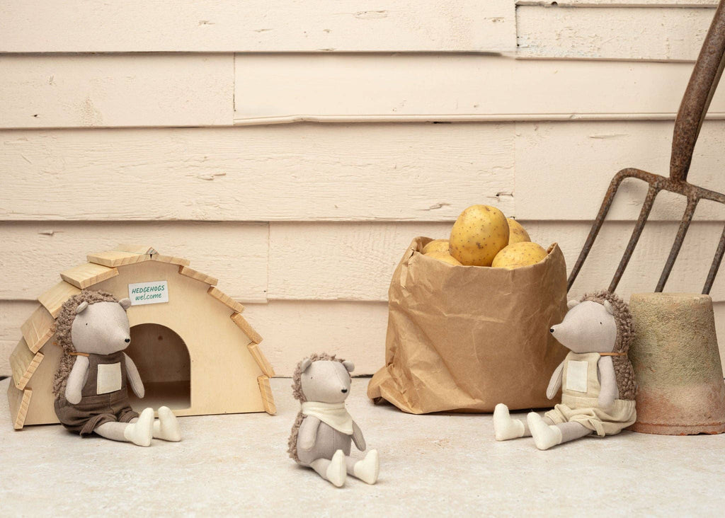 Three pocket-sized Poppet The Baby Hedgehog plush toys sit near a small wooden house labeled "Hedgehogs Welcome." A brown paper bag with potatoes and a garden fork, ideal for gardening, rest nearby against a light wooden wall.