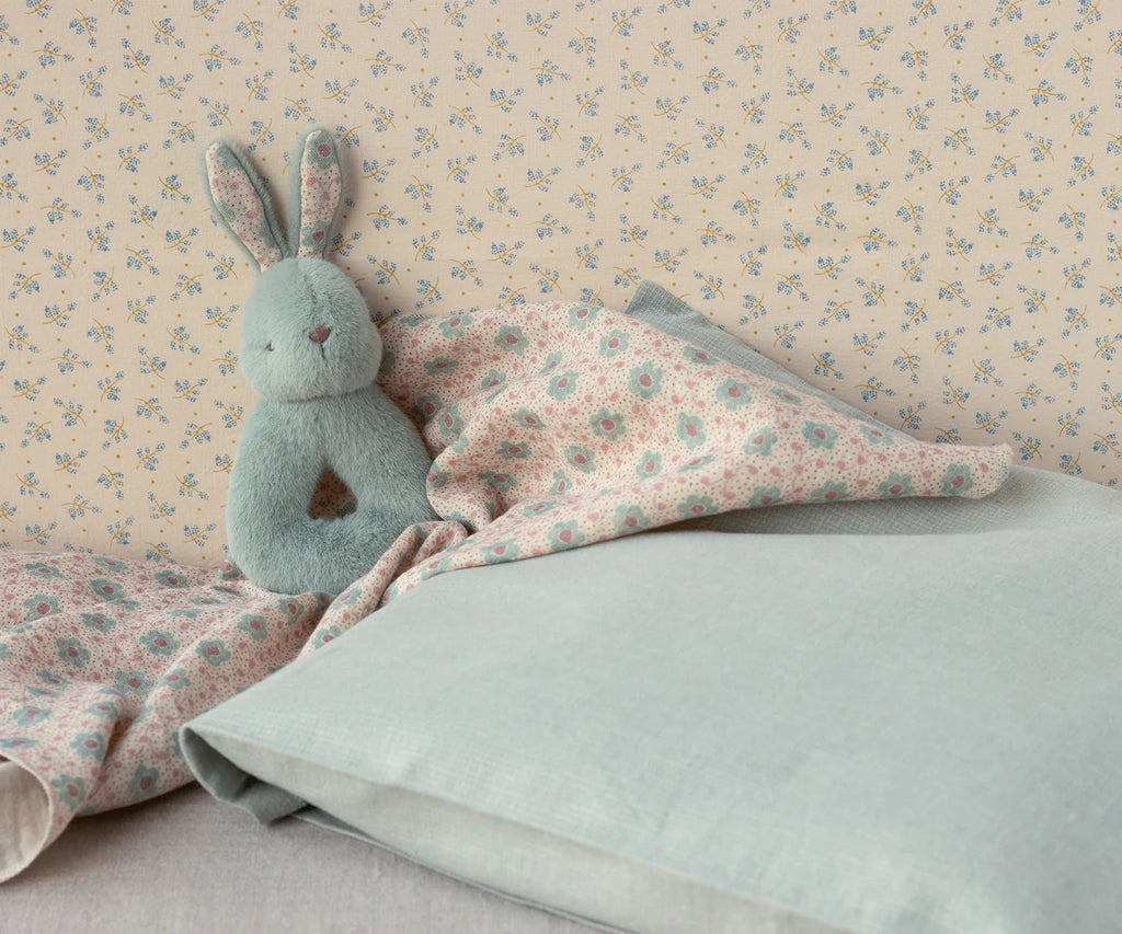 The Maileg Bunny Rattle Set - Mint rests on pastel floral bedding, complemented by wallpaper with delicate blue flowers. Made from soft cotton in pink and blue tones, the bedding creates a cozy and serene ambiance.