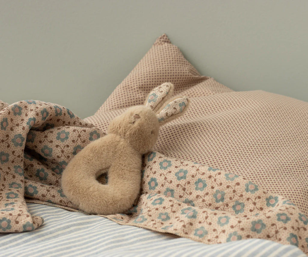 A Maileg Bunny Rattle Set in Cream Peach sits against two pillows on a bed. The bedding features a brown and blue floral pattern, while the pillows have a subtle geometric design. A light, neutral background adds to the cozy ambiance.