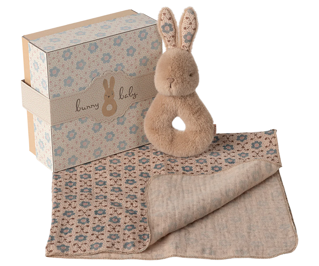 The Maileg Bunny Rattle Set - Cream Peach includes a plush bunny toy with a ring handle and charming rattle, paired with a partially rolled, floral-patterned baby blanket. A decorative gift box with matching floral designs and the phrase "bunny baby" completes this delightful set.