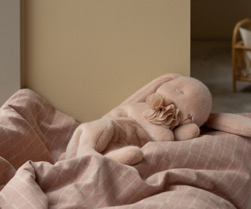 A Maileg Plush Sleeping Bunny, with droopy ears and a fabric rose on its chest, rests on a pink checkered blanket against a light yellow wall. A chair softly blurs in the background, enhancing the cozy atmosphere. This charming bunny ships in one week.