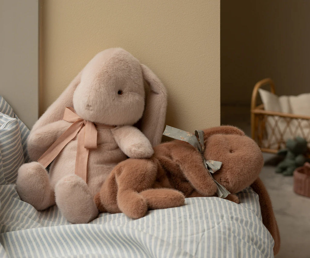 Two Maileg plush bunny toys rest on a striped bed against a light wall. The soft brown one, crafted from Maileg cotton fabric, wears a polka dot bow. Nearby, the light pink bunny dozes peacefully with a wicker basket and other sleeping bunny toys in the dim background.