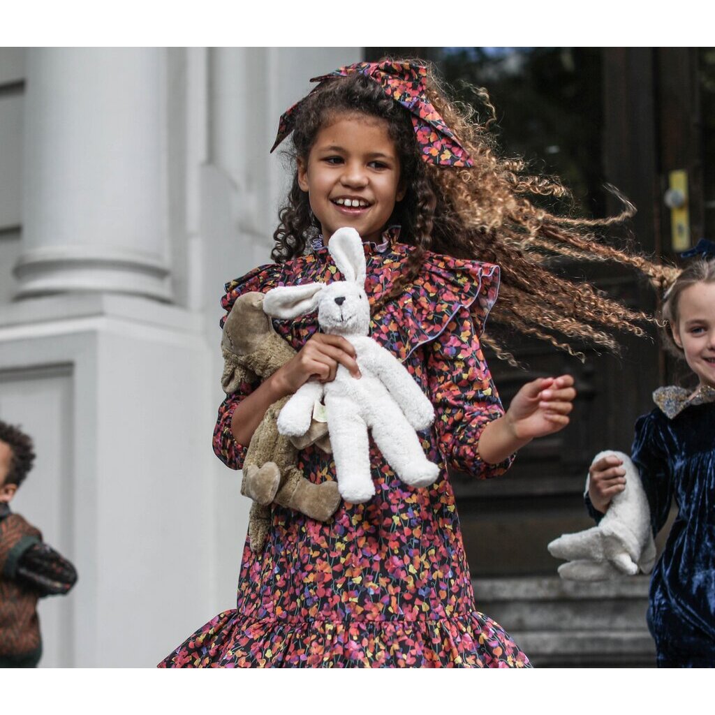 A smiling young girl in a colorful floral dress holds the Senger Naturwelt Stuffed Animal - White Bunny, handmade in Germany from organically grown cotton, while standing outside with other children as her curly hair blows in the wind.