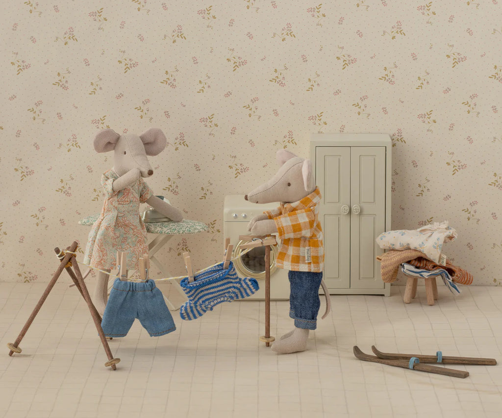 Two mouse dolls are in a cozy laundry scene; one in a dress irons clothes, while the other, wearing a Maileg Knitted Sweater and Pants for Big Brother Mouse (ships in one week), hangs them on a rack. A cabinet, stool with clothes, and skiing gear complete the setting with touches of blue stripes on some garments.