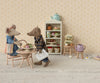 Dad Mouse, in denim pants and holding a shopping bag, enjoys tea and pastries at a tiny table with ingredients. Floral wallpaper sets a cozy background with a shelf of more pastries and teapots. The Maileg Dad Mouse (Light Brown) ships in one week.