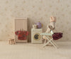 A charming scene features a toy mouse dressed in a Maileg Skirt and Knitted Blouse For Mum Mouse, ironing clothes. Nearby are a small washing machine, wardrobe, colorful teapot, and stool with a knitted blouse against floral wallpaper. (ships in one week).