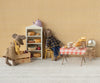 In a delightful miniature bakery scene, a stuffed mouse wearing the Maileg Dress and Bag for Mum Mouse holds a wooden box, while another in overalls reaches for bread. A table with pastries, a cheese hole bag, and a teapot completes the setting against patterned wallpaper.