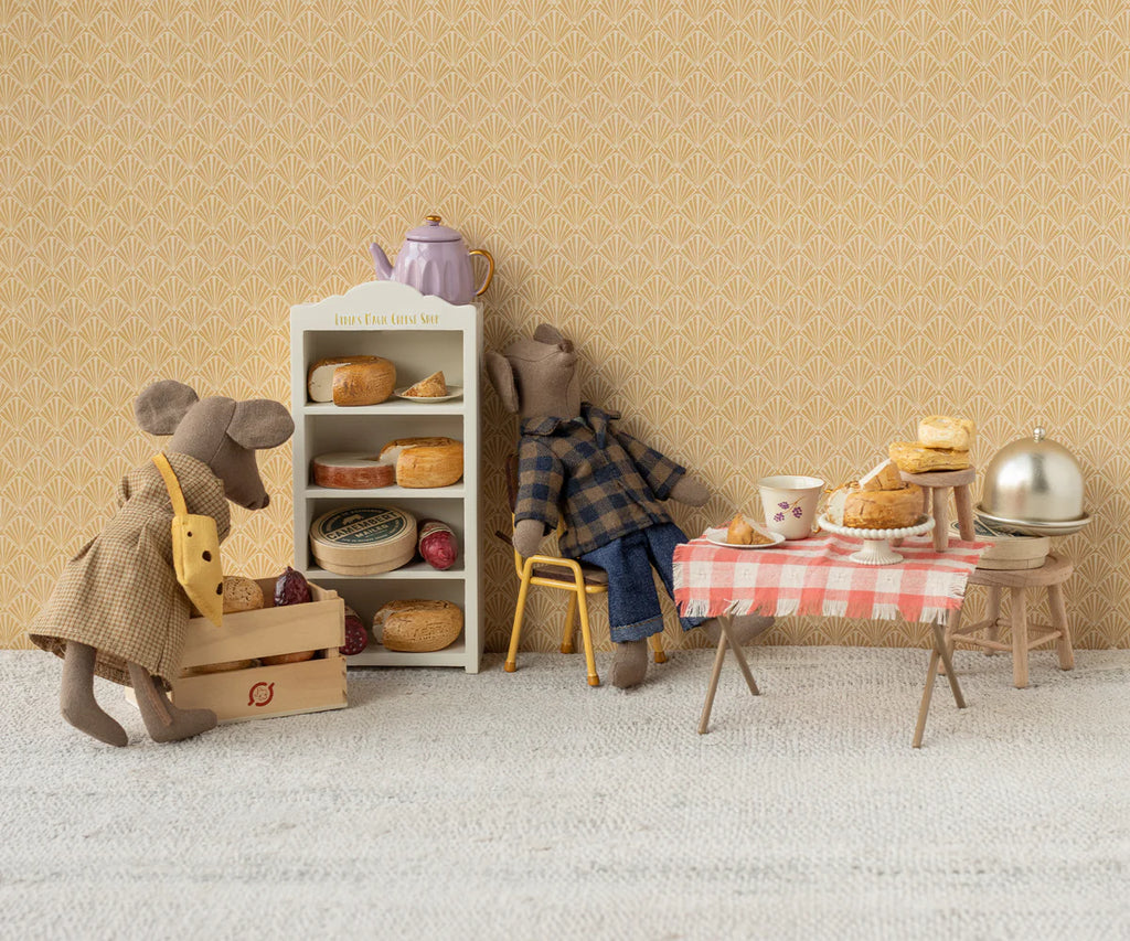In a delightful miniature bakery scene, a stuffed mouse wearing the Maileg Dress and Bag for Mum Mouse holds a wooden box, while another in overalls reaches for bread. A table with pastries, a cheese hole bag, and a teapot completes the setting against patterned wallpaper.