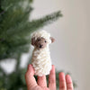 A hand holds the Fluffy Sheep Finger Puppet, a small knitted creation handmade in Nepal, in front of a blurred green foliage background. The cream-colored sheep, crafted from soft, chunky yarn, features detailed brown facial accents.
