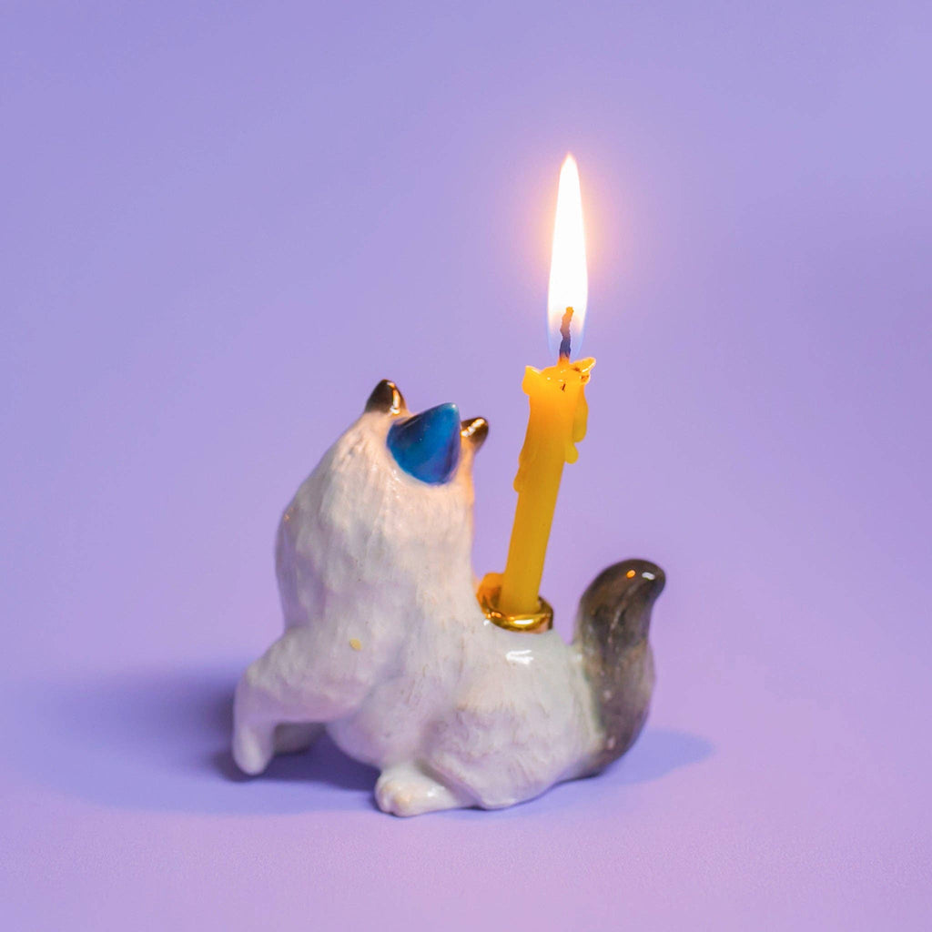A ceramic cat-shaped candle holder, similar to a Ragdoll Cat Cake Topper, features a yellow candle glowing brightly against a lavender background. The white feline with gray tail and ears faces away, accentuating the cheerful flicker—ideal for cat-themed parties.