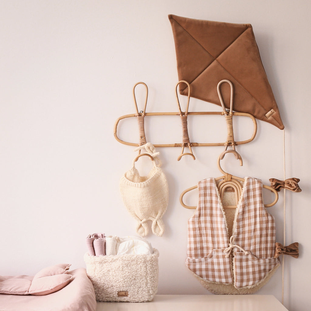 A decorative wall rack in a children's room holds items including a Velvet Kite Wall Decor - Brown, a woven backpack, and two checkered vests. Below, there is a plush basket with folded clothes. A light pink bedding set, OekoTex® Standard 100 certified, is partially visible on the left side. The colors are soft and neutral.