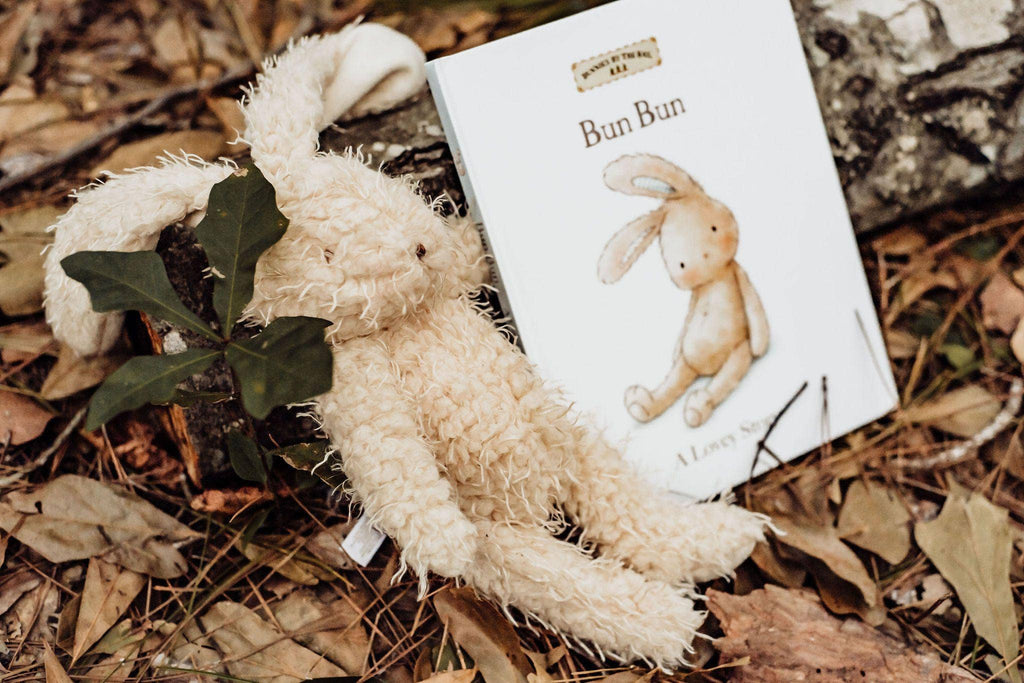 Harey the Bunny, a plush toy with embroidered details, rests on fallen leaves beside a book titled "Bun Bun," featuring a similar illustration. The scene has a vintage vibe with surrounding greenery and a tree trunk in the backdrop.