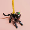 A lifelike black cat-shaped candle holder, wearing a striped party hat, holds a tall yellow candle on a light pink surface—ideal for cat-themed parties.
