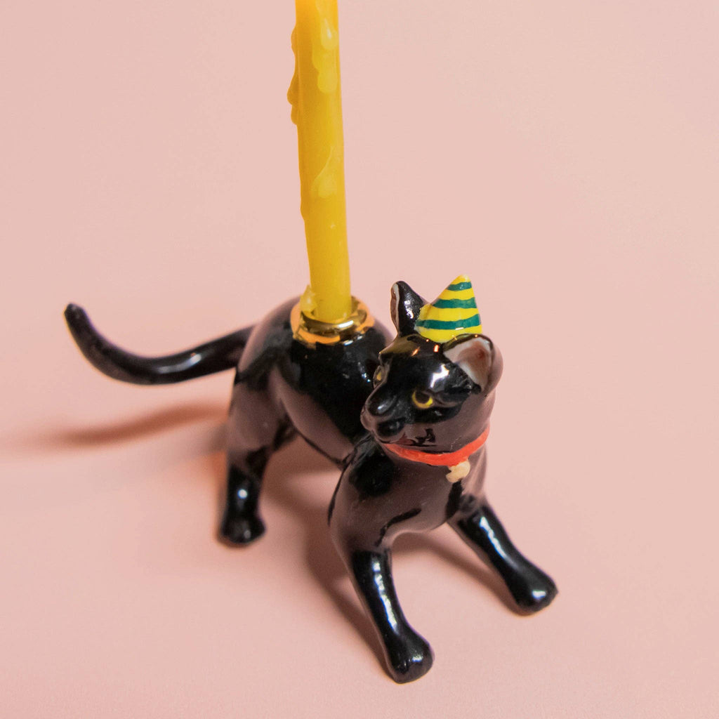 A lifelike black cat-shaped candle holder, wearing a striped party hat, holds a tall yellow candle on a light pink surface—ideal for cat-themed parties.