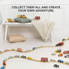 A playroom with a wooden train set on light wood flooring, colorful Classic Wooden Sports Cars, and FSC-certified wood buildings. A cushioned bench with pillows and a blanket is in the background. The text above reads, "Collect them all and create your own adventure.