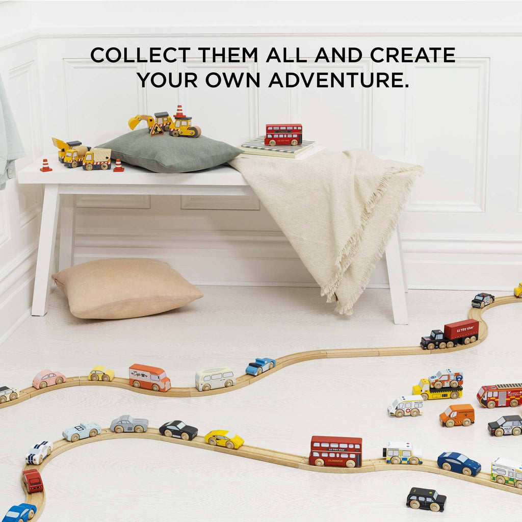 A playroom with a wooden train set on light wood flooring, colorful Classic Wooden Sports Cars, and FSC-certified wood buildings. A cushioned bench with pillows and a blanket is in the background. The text above reads, "Collect them all and create your own adventure.